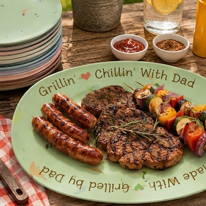 May include: A light green oval serving platter featuring the text "Grillin' Chillin' With Dad" and "Made With Grilled by Dad" with grilled sausages, steaks, and vegetable skewers. A stack of pastel plates, a metal spatula, and condiments are nearby.