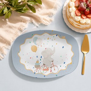 May include: An oval serving platter with a light blue border and a central white scalloped area featuring a cartoon elephant riding a red bicycle. The elephant carries stacked gift boxes and colourful balloons, surrounded by scattered confetti.