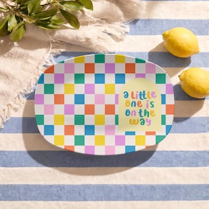 May include: An oval serving platter featuring a vibrant, multi-coloured checkerboard pattern in shades of orange, yellow, blue, pink, and green. A pale yellow arch shape on the right side displays the text "a little one is on the way".