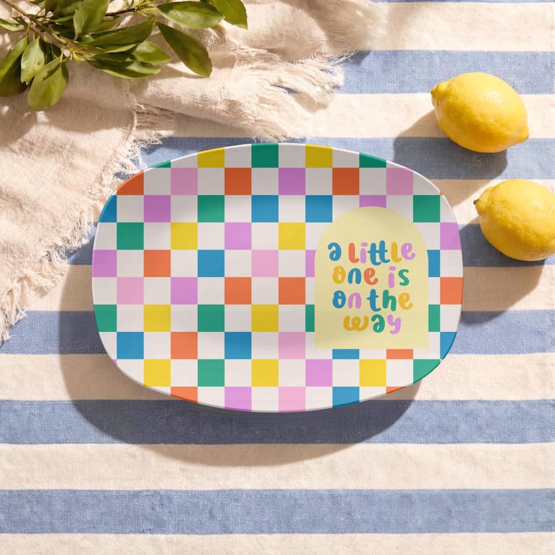 May include: An oval serving platter featuring a vibrant, multi-coloured checkerboard pattern in shades of orange, yellow, blue, pink, and green. A pale yellow arch shape on the right side displays the text "a little one is on the way".