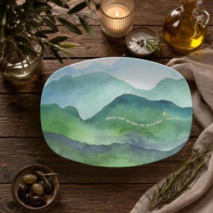 May include: An oval ceramic platter featuring a watercolour landscape of rolling mountains in shades of blue and green. The text "where God guides He provides" is printed in white script across the centre, accompanied by a small gold star.