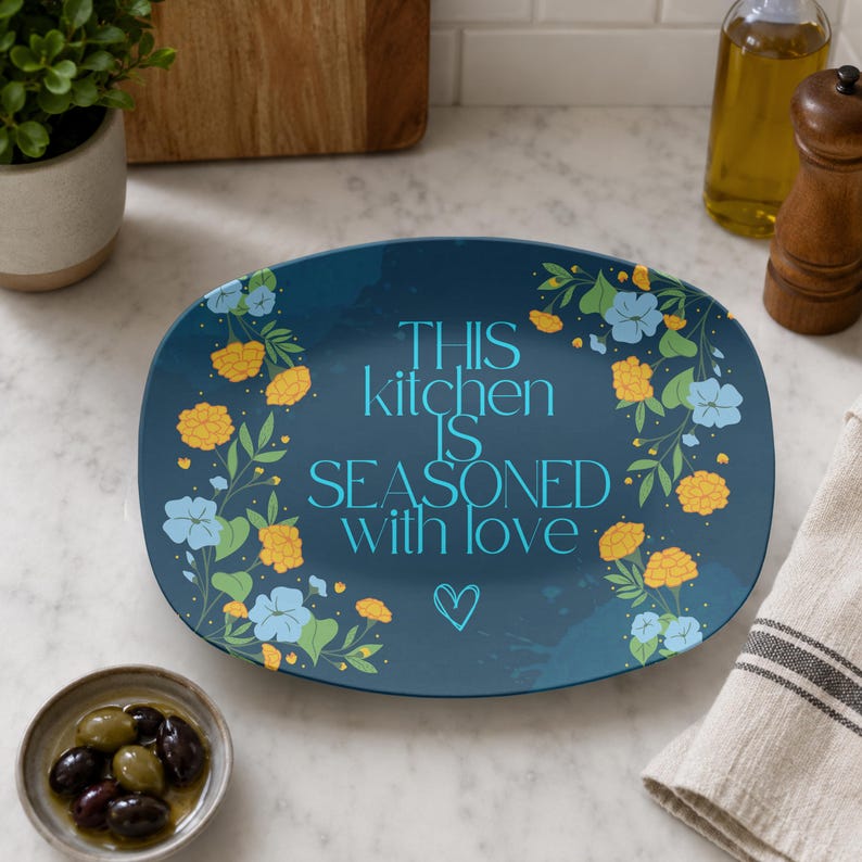 May include: An oval-shaped dark blue serving platter featuring a floral pattern of yellow and light blue flowers along the sides. The centre displays the text "THIS kitchen IS SEASONED with love" in light blue lettering, accompanied by a small heart icon.
