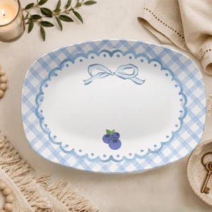 May include: An oval ceramic serving platter featuring a light blue and white gingham pattern border. The centre is white with a scalloped blue edge, a light blue ribbon bow illustration at the top, and two small blue blueberries at the bottom.