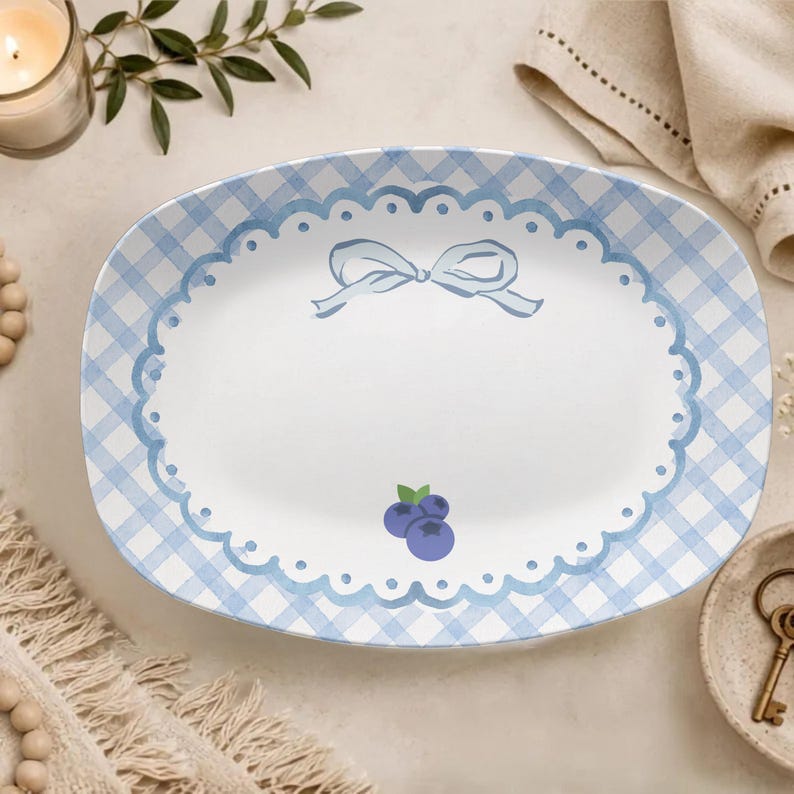 May include: An oval ceramic serving platter featuring a light blue and white gingham pattern border. The centre is white with a scalloped blue edge, a light blue ribbon bow illustration at the top, and two small blue blueberries at the bottom.