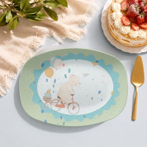 May include: An oval serving platter featuring a central illustration of a light-coloured bear riding a red bicycle with a crate of gifts on the back, accompanied by floating balloons, set against a pale green background with a scalloped blue border.