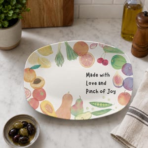 May include: An oval white ceramic serving platter decorated with watercolour illustrations of various fruits and vegetables including lemons, tomatoes, squash, and peas. The text "Made with Love and Pinch of Joy" is printed in black in the centre.