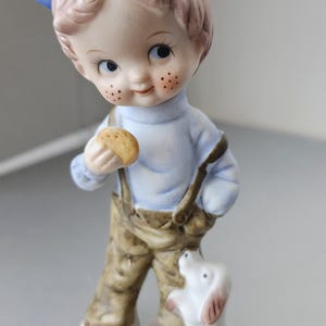 May include: A porcelain figurine of a child wearing a light blue shirt, brown trousers with suspenders, and a blue and white cap. The child holds a small round object in one hand, while a small white dog with brown spots stands at their side.