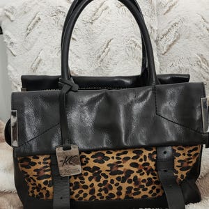 May include: A black leather handbag featuring a leopard print panel on the front, two top handles, and a silver-toned metal tag engraved with the letters KC. The bag includes two vertical leather straps and silver metal accents on the sides.