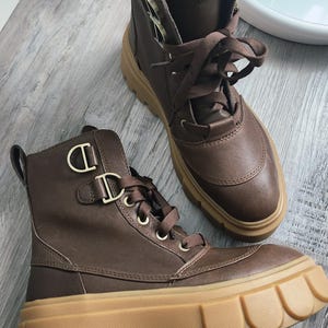 May include: A pair of brown leather ankle boots with a thick, tan-coloured lug sole. The boots feature brown laces, gold-toned metal eyelets, and a pull tab at the heel. The brand name SOREL is printed in white on a black tag on the tongue.