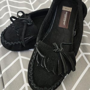 May include: A pair of black suede moccasins featuring a decorative fringe and a tied lace detail on the vamp. The interior footbed includes a brown rectangular label with the text Manitobah Mukluks and a size tag indicating LG/MG.