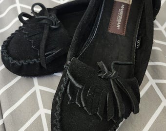 May include: A pair of black suede moccasins featuring a decorative fringe and a tied lace detail on the vamp. The interior footbed includes a brown rectangular label with the text Manitobah Mukluks and a size tag indicating LG/MG.