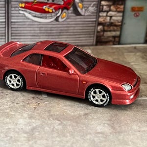 Könnte beinhalten: Ein metallisch rotes Druckguss-Spielzeugauto mit zweitüriger Coupé-Karosserie, Schiebedach und silbernen Sechsspeichenrädern mit weißer Yokohama-Beschriftung auf den schwarzen Reifen. Das Fahrzeug hat klare Fenster und einen Heckspoiler.