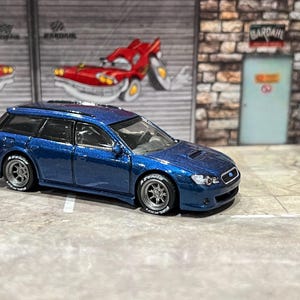 May include: A metallic blue die-cast station wagon toy car featuring a hood scoop, dark grey multi-spoke wheels, and tires with white lettering. The vehicle has a sleek body design with tinted windows and a rear spoiler, capturing the look of a performance-oriented estate car in a miniature scale.