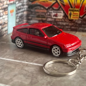 May include: A red die-cast model car keychain featuring a hatchback design with white six-spoke wheels, black trim, and Advan branding on the tires. The miniature vehicle is attached to a standard silver metal split ring by a short chain.