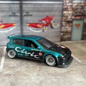 May include: A teal and black die-cast Honda Civic toy car featuring white 'Civic' lettering on the side, a rear spoiler, and silver wheels with white tire lettering. The car includes small red circular decals and Japanese text on the body.