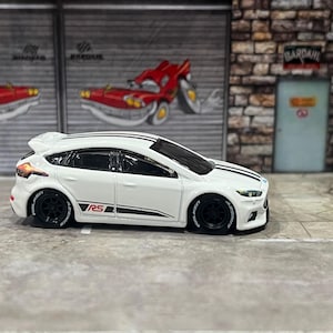 May include: A white die-cast Ford Focus RS toy car featuring black racing stripes on the hood and roof, black wheels with white lettering, and a red RS logo on the side door. The vehicle has a rear spoiler and tinted windows, presented in profile.