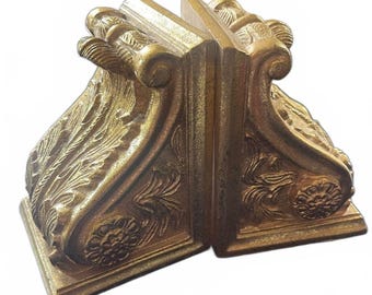 May include: A pair of gold-coloured bookends featuring ornate, raised relief carvings of acanthus leaves and circular floral motifs. Each bookend has a curved, scroll-like top edge and a rectangular base, showcasing a textured, metallic finish.