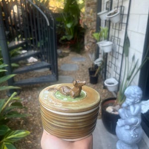 May include: A round ceramic lidded jar with a brown, ribbed exterior and a smooth, glossy finish. The lid features a small, sculpted brown dog figurine resting on a green, circular base with white floral accents, centred on the top surface.