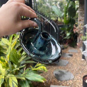 May include: A dark teal and blue glazed ceramic candle holder with a circular base, a central cylindrical socket for a candle, and a curved handle attached to the rim, featuring a glossy finish with subtle variations in colour throughout the glaze.