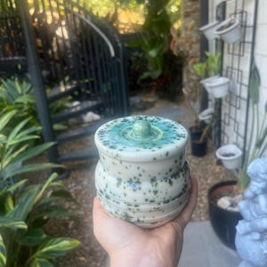 May include: A small, round ceramic jar with a lid, featuring a glossy white base glaze speckled with dark green and black spots. The lid has a central knob and a gradient of teal and green glaze that transitions into the white speckled body.