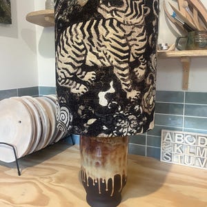 Könnte beinhalten: Eine zylindrische Tischlampe mit einem schwarz-beigen Stoffschirm mit wiederkehrendem Tiger- und Blumenmuster. Der Fuß ist ein Keramikgefäß mit einer glatten, glänzenden Glasur, die von einem hellen Beige oben zu einem Dunkelbraun unten verläuft.