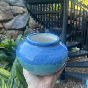 May include: A small, round ceramic vase featuring a two-tone glaze with a vibrant cobalt blue upper section that transitions into a lighter turquoise blue base. The vase has a wide, smooth opening with a thick rim and a glossy, reflective finish.