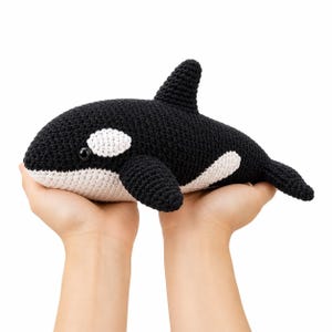 May include: A crochet orca toy featuring a black and white yarn pattern. The stuffed animal has a prominent dorsal fin, a rounded head with a black plastic eye, and a white patch near the eye. The body is crafted with a textured stitch pattern.
