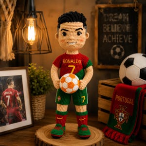 Puede incluir: Una muñeca de ganchillo hecha a mano de un jugador de fútbol con pelo negro, vistiendo una camiseta roja y verde con el nombre RONALDO y el número 7, pantalones cortos verdes con el número 7 y calcetines rojos, sosteniendo un balón de ganchillo.