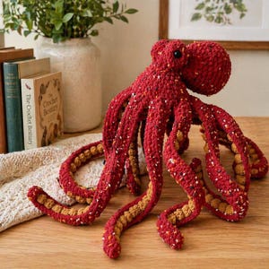 May include: A handmade crochet octopus toy featuring a deep red yarn body with speckled white and black flecks. The underside of the eight curled tentacles is a contrasting tan color with a textured, bumpy surface. It has two black eyes.