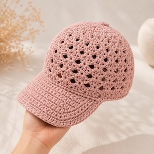 Puede incluir: Una gorra de béisbol de color rosa empolvado hecha de hilo de ganchillo. La corona presenta un intrincado patrón calado en forma de estrella, mientras que la visera está construida con un punto de ganchillo denso y sólido con un nudo central.