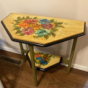 May include: A two-tiered semi-circular side table with a light yellow finish and black edges. Both the top and lower shelves feature hand-painted floral arrangements in shades of red, orange, purple, and blue, supported by four green legs.