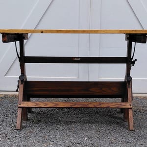 May include: A drafting table with a light-colored rectangular wooden top and a dark-stained wooden base. The structure features adjustable metal hardware, including side knobs and curved supports, with a horizontal footrest near the bottom.