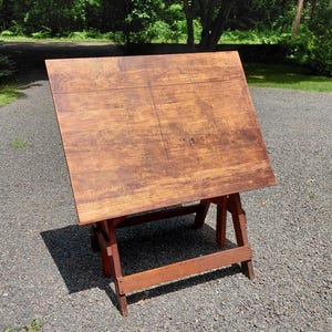 May include: A wooden drafting table with a large, rectangular, tilted work surface featuring a visible wood grain pattern. The table is supported by a sturdy, four-legged frame with horizontal crossbars, all finished in a warm, medium-brown stain.
