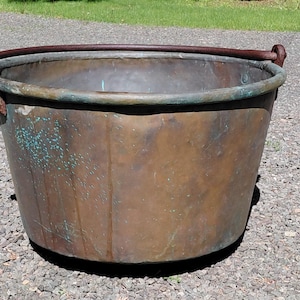 May include: A large, antique copper bucket with a dark brown handle and aged patina. The bucket has a wide opening and a rounded bottom. The copper has areas of green oxidation, adding to its vintage appeal. Ideal for decorative use or as a rustic accent.