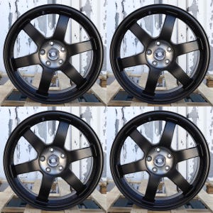 May include: A set of four matte black six-spoke alloy wheels featuring a circular center hub with five lug nut holes. Each wheel rim displays the embossed text "FORGED" and "MADE IN JAPAN" along the outer edge of the spoke structure.