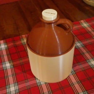 May include: A brown ceramic jug with a rounded body and a handle, featuring a smooth cream-colored cylindrical base. The top of the jug is fitted with a white circular stopper that has a thin horizontal slot cut into the center of its surface.