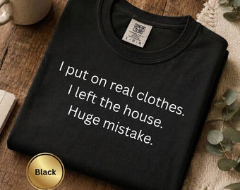 May include: A black crew neck t-shirt featuring the white printed text: I put on real clothes. I left the house. Huge mistake. The shirt has a Comfort Colors brand label on the inside collar indicating a size large and is made in Honduras.