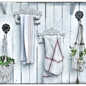 Könnte beinhalten: Weiße bemalte Holzwand mit drei dekorativen Haken. An zwei Haken hängen weiße Handtücher, an dem dritten Haken hängt ein weißer gehäkelter Hängepflanztopf mit grünen Pflanzen.
