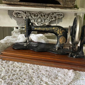 Könnte beinhalten: Antike Singer Nähmaschine, schwarz mit goldenen Blumenmustern, auf einem Holzsockel. Sie hat ein großes silbernes Schwungrad und ein dekoratives Emblem. Ein klassisches Stück für Nähprojekte.