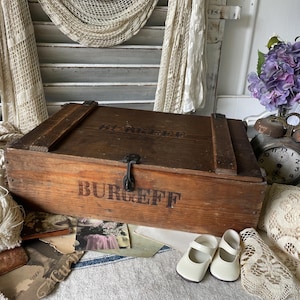 Könnte beinhalten: Eine alte Holzkiste mit dem Aufdruck "BURGEFF". Das Bild zeigt auch eine Vintage-Uhr, Spitze und ein Paar weiße Schuhe, was eine nostalgische Atmosphäre erzeugt.