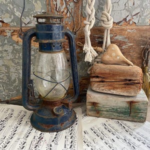 May include: A vintage blue lantern with a clear glass globe, set on a sheet of musical notes. The lantern has a handle and a rusty appearance. Beside it are wooden blocks, a rope, and a wooden heart with white floral accents.
