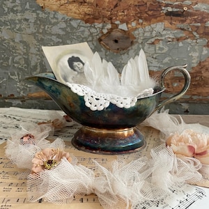 May include: A vintage silver gravy boat with a blue and gold patina, holding white feathers and a small framed photograph. The boat rests on sheet music, with lace and fabric flowers.