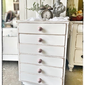 Peut inclure: Une commode blanche peinte avec six tiroirs et des boutons en céramique rose. La commode a une finition vieillie et est posée sur un sol en bois.