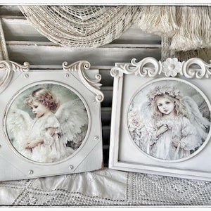 May include: Two square white wooden frames featuring circular cutouts. Each frame displays a print of a young angel with wings. The left frame has decorative scrollwork corners and exposed screw heads. The right frame features a floral carved top.