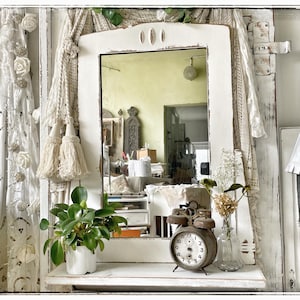 Op de afbeelding: Een wit omlijste spiegel met een verweerde afwerking is het middelpunt, omringd door witte kanten gordijnen en decoratieve kwasten. Een kleine potplant, een vintage wekker en een glazen vaas met gedroogde bloemen staan op een plank.