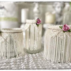 Könnte beinhalten: Drei Glasvasen, verziert mit weißer Häkelspitze und rosa Stoffblumen. Die Vasen stehen auf einer weißen Häkeltischdecke.