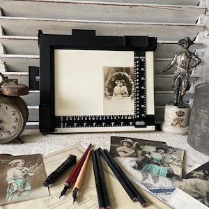 May include: An antique still life featuring a vintage alarm clock, a black and white photo, and a black photo enlarger. A small statue and pencils are also present. The photo enlarger has a ruler with inch measurements.