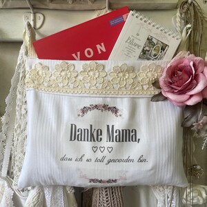 May include: A white fabric pocket with a cream lace trim, holding a red card and a spiral-bound notebook. The pocket has the text "Danke Mama" and a floral design. A large pink rose is attached to the side.