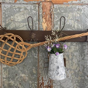 Könnte beinhalten: Ein Wandgarderobe im Vintage-Stil mit einem gewebten Teppichklopfer, einem gewebten Besen und einer dekorativen Metallgießkanne mit lila Blumen. Die Garderobe ist aus dunklem Holz gefertigt und hat mehrere Metallhaken.