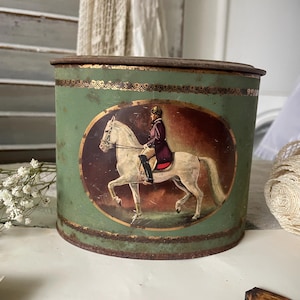 Può includere: Scatola di metallo verde vintage con coperchio. La scatola presenta un'immagine ovale di una persona su un cavallo bianco. L'immagine è incorniciata in oro. La scatola ha un aspetto arrugginito e invecchiato.